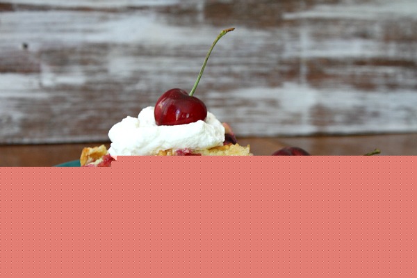 serving of Easy Cherry Cobbler on a green plate set on a wooden table. Garnished with whipped cream and fresh cherries with stems