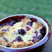 cherry cobbler in a round burgundy casserole dish sitting on a wooden railing with green background