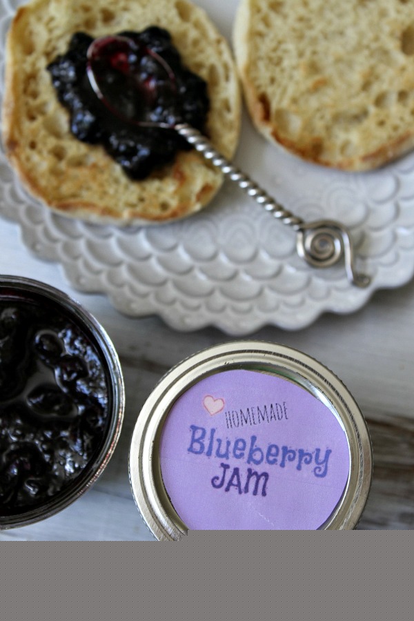 overhead shot of blueberry jam in a jar with the lid displayed too, The lid has a purple label. english muffin with spoon of jam on it on a plate.