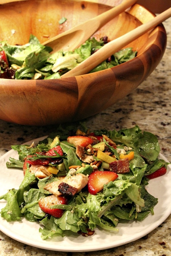 serving of strawberry chicken salad on a white pate with salad in a big wooden bowl with wooden serving spoons in the background