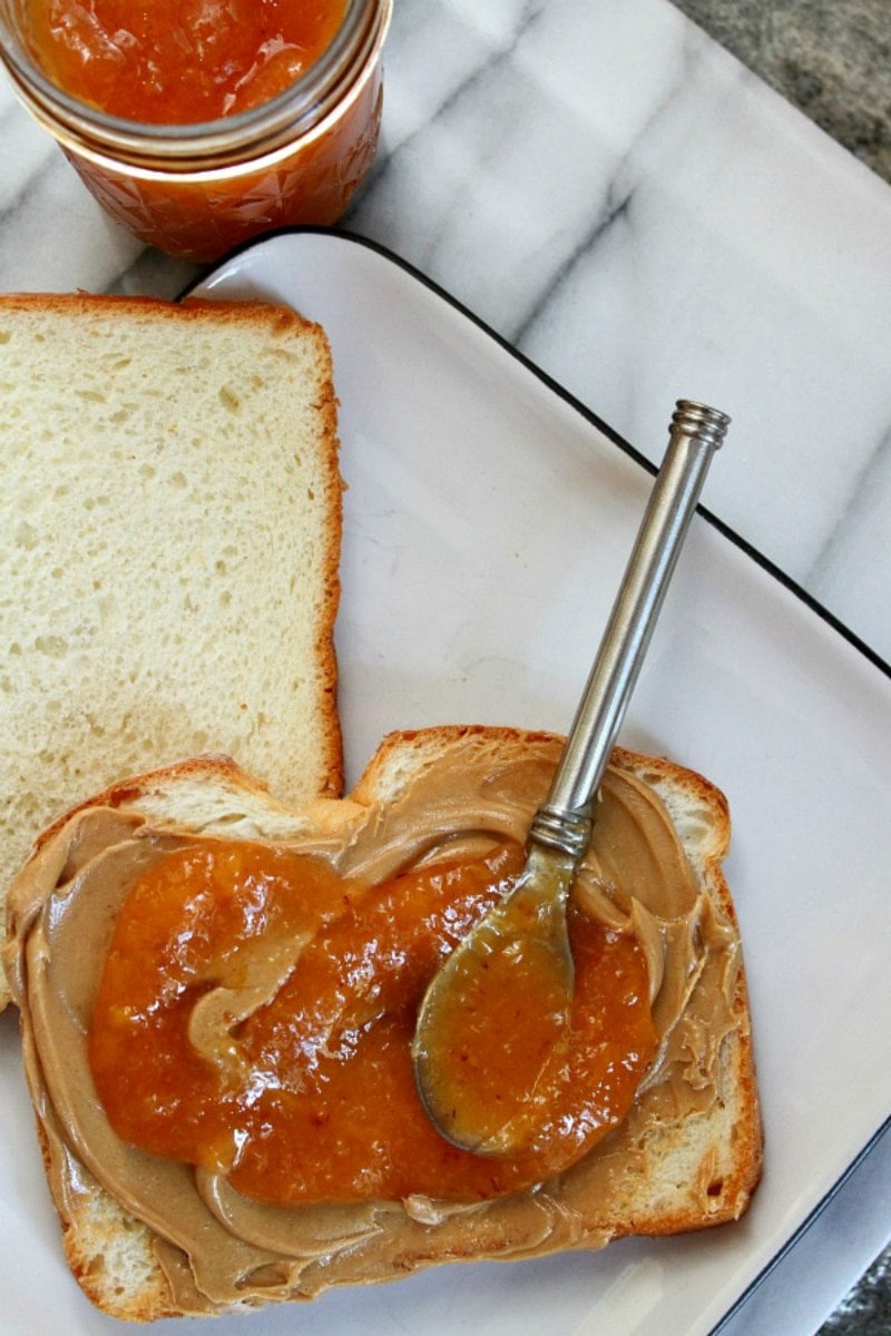 bread with peanut butter and peach jam with a spoon resting on the bread. jar of peach jam in background