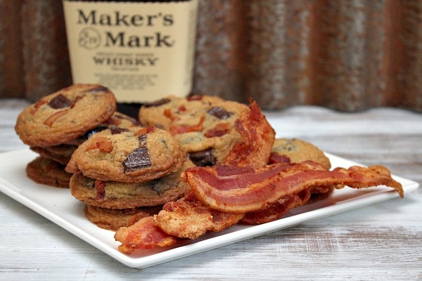 Bourbon Bacon Chocolate Chunk Cookies sitting on a white plate with some bacon slices. Maker's Mark Whisky in the background