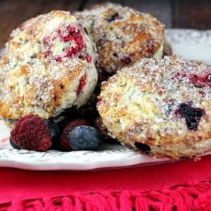 three berry scones on a white platter with fresh berries scattered. Pink cloth napkin underneath