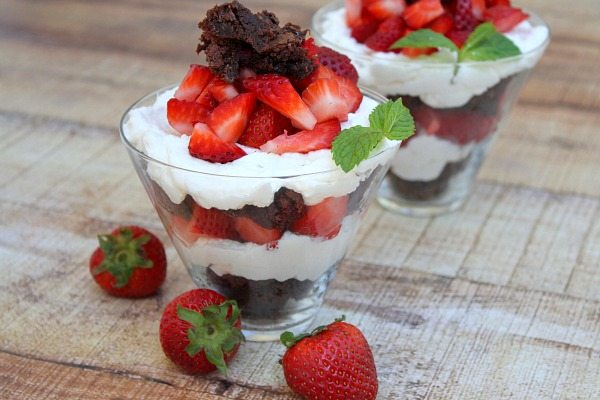 two Brownie Strawberry Shortcakes in clear glasses with strawberries scattered around
