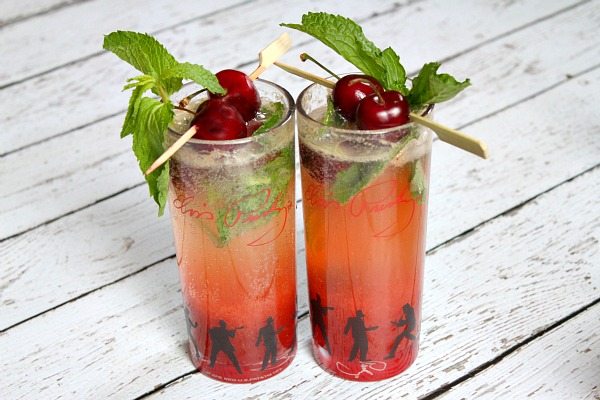 two cherry mojitos displayed on a white wood plank board garnished with fresh cherries and mint