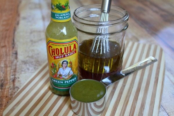 Making dressing for salad with a bottle of cholulua green hot sauce, a measuring cup and a jar with a whisk. set on a cutting board.