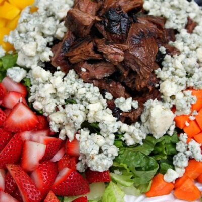 grilled steak summer salad on a white plate with steak piled in the middle surrounded by blue cheese and vegetables and fruit