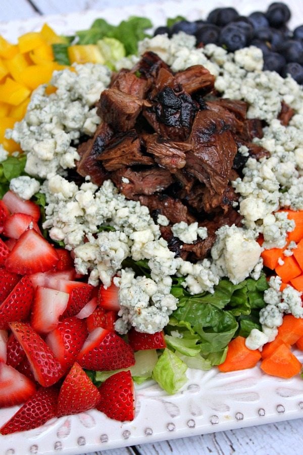 Grilled Steak Chopped Summer Salad on a square white plate with steak piled in the middle and veggies, lettuce and fruit and blue cheese crumbles piled around the steak. on a white plank table