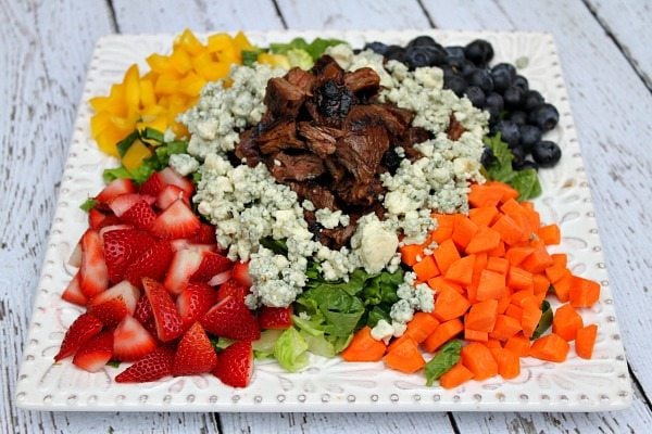 Grilled Steak Chopped Summer Salad on a square white plate with steak piled in the middle and veggies, lettuce and fruit and blue cheese crumbles piled around the steak. on a white plank table