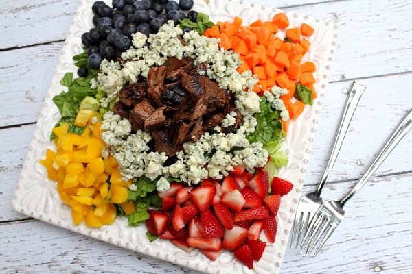 Grilled Steak Chopped Summer Salad on a square white plate with steak piled in the middle and veggies, lettuce and fruit and blue cheese crumbles piled around the steak. two forks on the side. on a white plank table