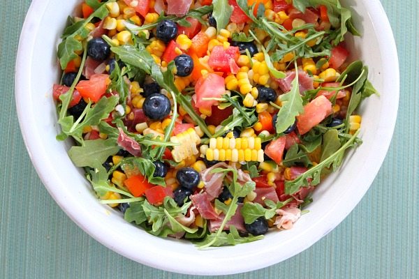 overhead shot of blueberry corn salad with prosciutto in a white bowl on a green background