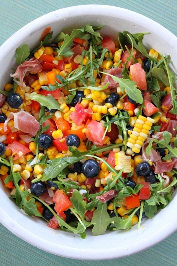 overhead shot of blueberry corn salad with prosciutto in a white bowl on a green background