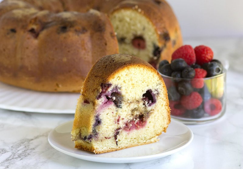 slice of two berry coffee cake on a white plate with the rest of the cake and bowl of berries in the background