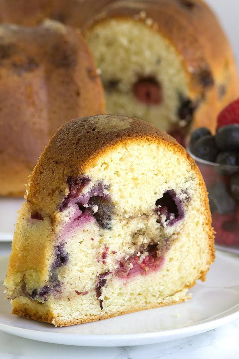 slice of two berry coffee cake on a white plate in front of the rest of the coffee cake