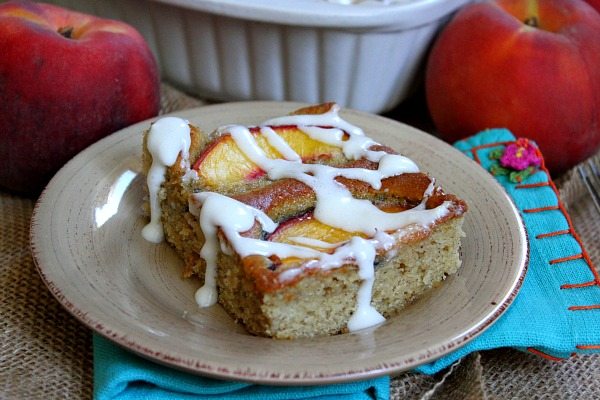 slice of fresh peach cake on a beige plate set on a turquoise napkin with a peach in the background and cake dish in background