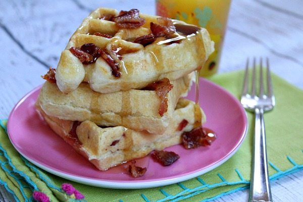 stack of three maple bacon waffles with more syrup and bacon on pink plate