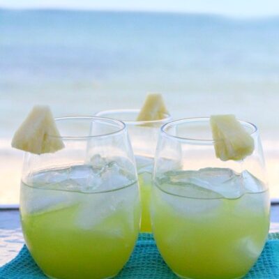 two pearl harbor cocktails sitting side by side garnished with pineapple sitting on a blue towel with the ocean in the background