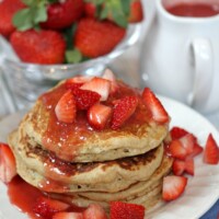 stack of strawberry ricotta pancakes. lots of fresh strawberries and strawberry sauce on top with fresh strawberries and pitcher of sauce in background.