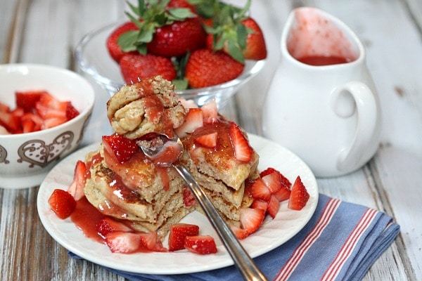 stack of strawberry ricotta pancakes with a fork taking a bite of them. lots of fresh strawberries and strawberry sauce on top with fresh strawberries and pitcher of sauce in background.