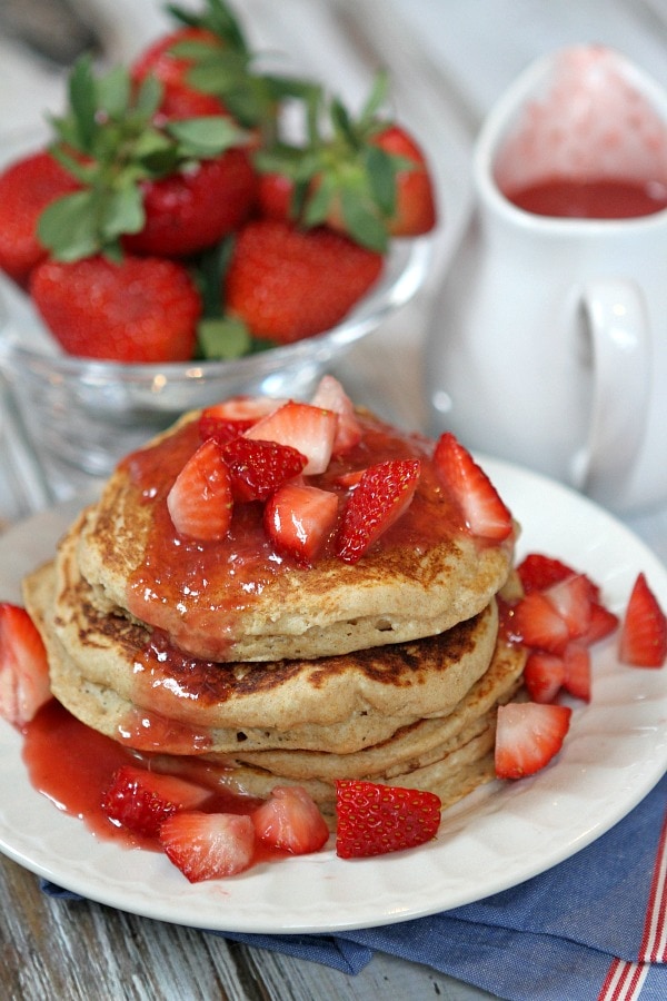 stack of strawberry ricotta pancakes. lots of fresh strawberries and strawberry sauce on top with fresh strawberries and pitcher of sauce in background.