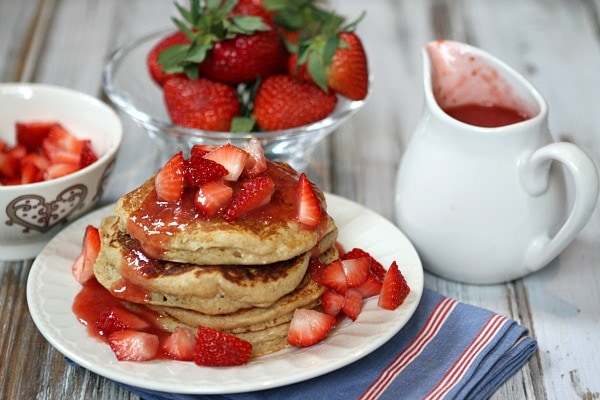 stack of strawberry ricotta pancakes. lots of fresh strawberries and strawberry sauce on top with fresh strawberries and pitcher of sauce in background.