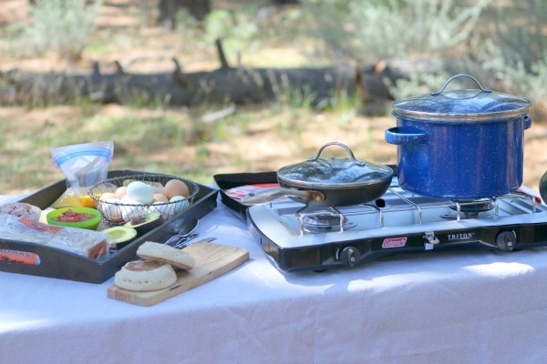 Set up at a campground for making Camping Breakfast Sandwiches. camp stove with blue pot and skillet on top. tray with ingredients for the sandwiches on top. english muffins on a cutting board.