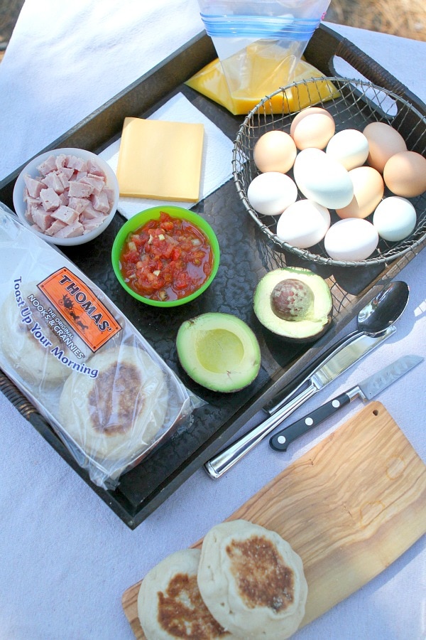 Ingredients displayed for Camping Breakfast Sandwiches: English muffins, avocado, salsa, ham, cheese, scrambled eggs in a plastic bag and eggs in a basket. Utensils and a cutting board on the side.