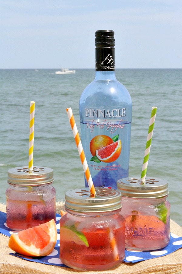 three ruby sunset cocktails in jars with straws and fresh grapefruit with a bottle of grapefruit vodka and the ocean in the background