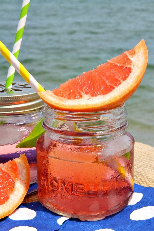 ruby sunset cocktail in a small glass jar garnished with fresh grapefruit, another cocktail next to it. Sitting on a blue napkin with white polka dots with the ocean in the background