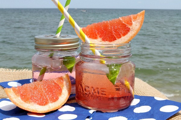 ruby sunset cocktail in a small glass jar garnished with fresh grapefruit, another cocktail next to it. Sitting on a blue napkin with white polka dots with the ocean in the background