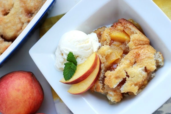 overhead shot of a serving of peach cobbler in a white serving dish garnished with ice cream, fresh peaches and mint leaves