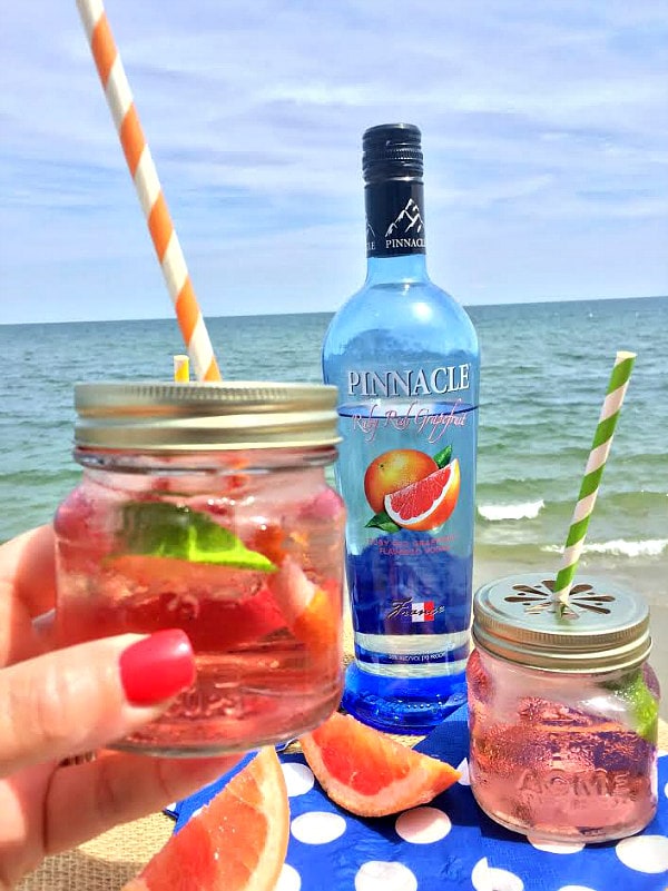 hand holding ruby sunset cocktail in a small glass jar with a red/white striped straw, bottle of vodka in the background and another cocktail, set on a blue napkin with white polka dots and the ocean in the background