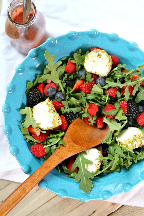 overhead shot of arugula berry and goat cheese salad in a turquoise bow with a wooden spoon inside and a bottle of salad dressing on the sidel set on a white cloth napkin on a wood board