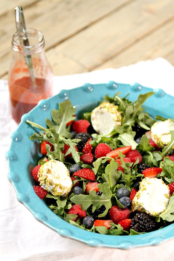 arugula berry and goat cheese salad in a turquoise bow with a wooden spoon inside and a bottle of salad dressing on the sidel set on a white cloth napkin on a wood board