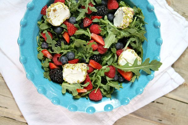 overhead shot of arugula berry and goat cheese salad in a turquoise bowl set on a white cloth napkin on a wood board