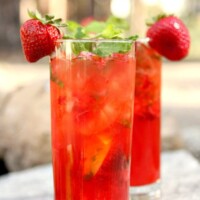 two strawberry mojitos in tall glasses garnished with fresh strawberries sitting on a rock in the backyard