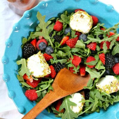 overhead shot of arugula berry and goat cheese salad in a blue bowl with a wooden serving spoon and a bottle of dressing on the side
