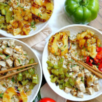 overhead shot of 3 white bowls filled with grilled chicken and pineapple rice bowls with chopsticks. Set on a white napkin with a wood board background with fresh bell peppers and green onions garnish.