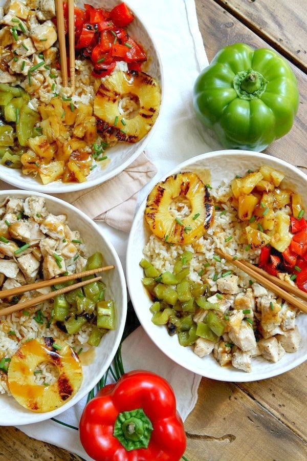 overhead shot of 3 white bowls filled with grilled chicken and pineapple rice bowls with chopsticks. Set on a white napkin with a wood board background with fresh bell peppers and green onions garnish.