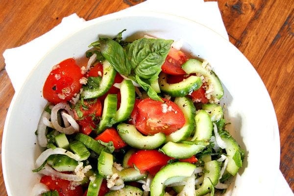 Overhead shot of Tomato Cucumber and Basil Salad in a white bowl with a white napkin on a wood surface