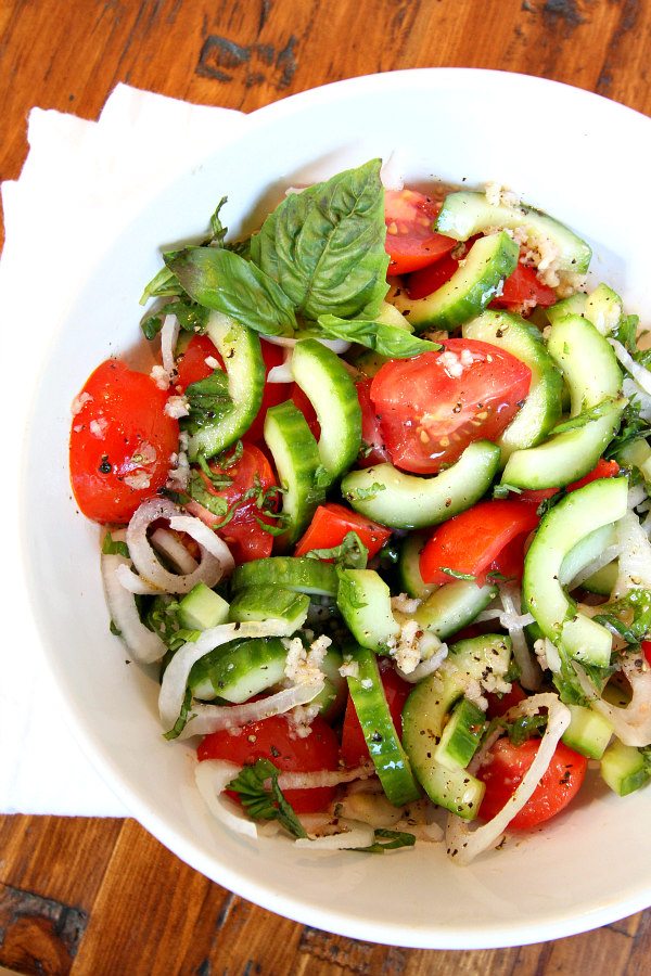 Overhead shot of Tomato Cucumber and Basil Salad in a white bowl with a white napkin on a wood surface