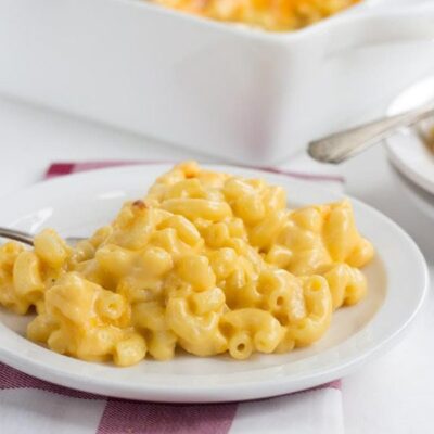 macaroni and cheese on a white plate set on a red and white plaid napkin with white casserole dish of macaroni and cheese in the background