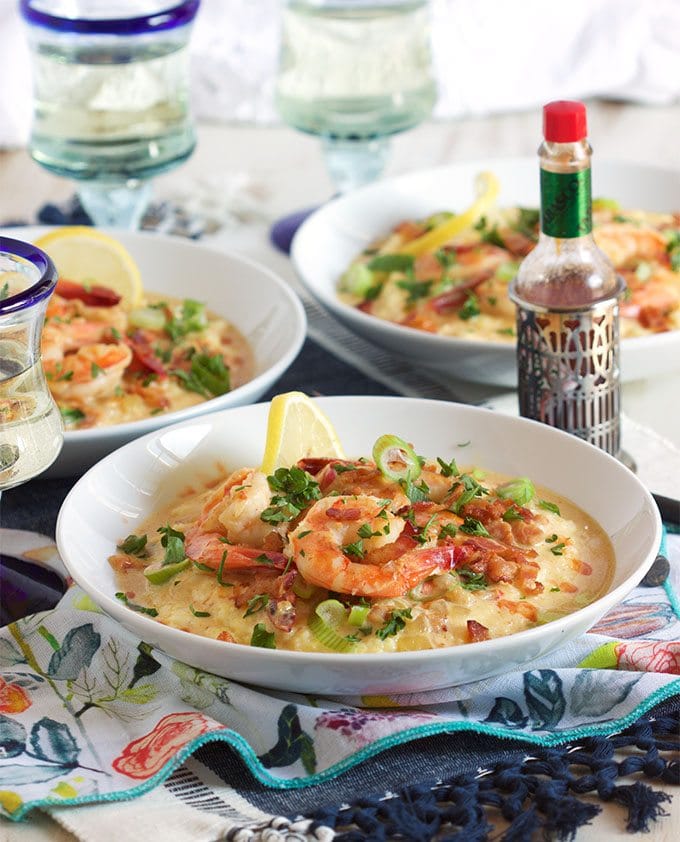 shrimp and grits in three white bowls on a table setting with patterned napkin and a bottle of hot sauce