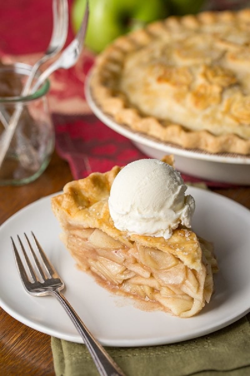 slice of apple pie on a plate with rest of pie in background