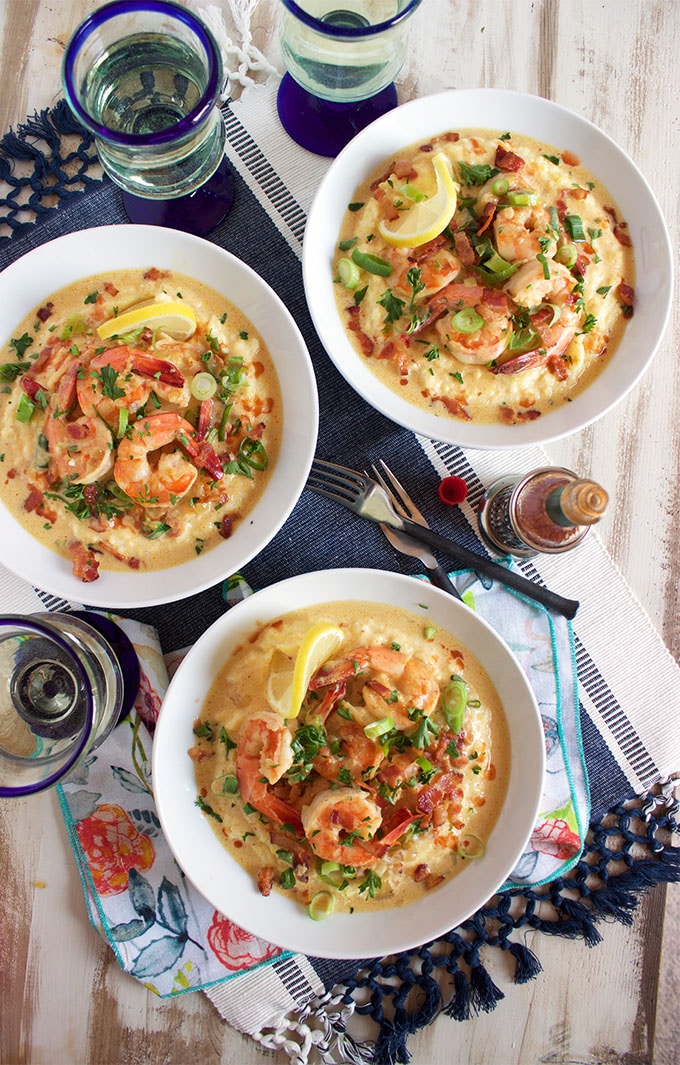 overhead shot of shrimp and grits in three white bowls with glasses and patterned napkin