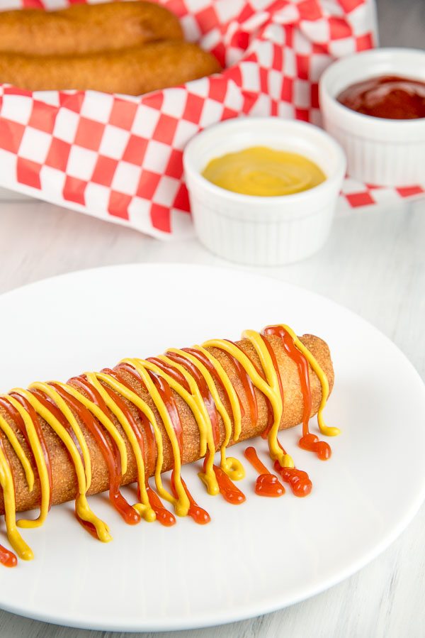 corn dog on a white plate drizzled with ketchup and mustard. basket of corn dogs with white/red checked paper and white dishes with ketchup and mustard in the background