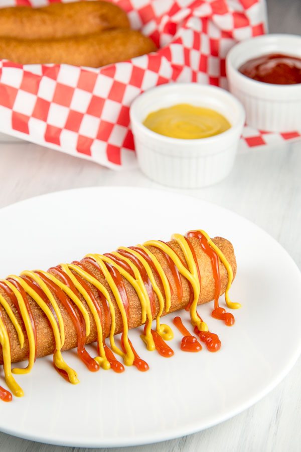 Make your own homemade corn dogs recipe from RecipeGirl.com #homemade #DIY #corn #dogs #corndogs #recipe #RecipeGirl corn dog on a white plate drizzled with ketchup and mustard.