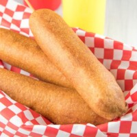 three Corn Dogs in a basket lined with red/white checked paper with ketchup and mustard bottles and dishes with mustard in it on the side