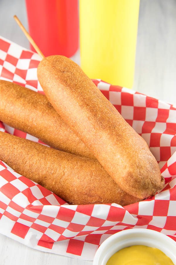 Three corn dogs in a red/white checkered paper lined basket with ketchup and mustard bottles in the background and a white dish with mustard in it in front