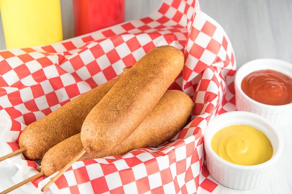 Three Corn Dogs in a basket with red/white checked paper with mustard and ketchup on the side in little white dishes